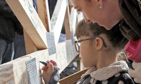 a woman watches a young girl write on a wooden beam with a marker