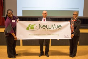 2 women and a man hold a banner with the NewVue Communities logo indoors with a projector screen partially visible in the background