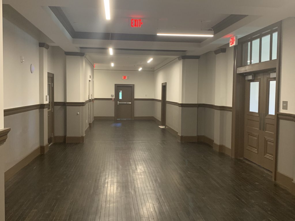 a hallway with dark wood floor, beige walls, dark brown doors, and red exit lights