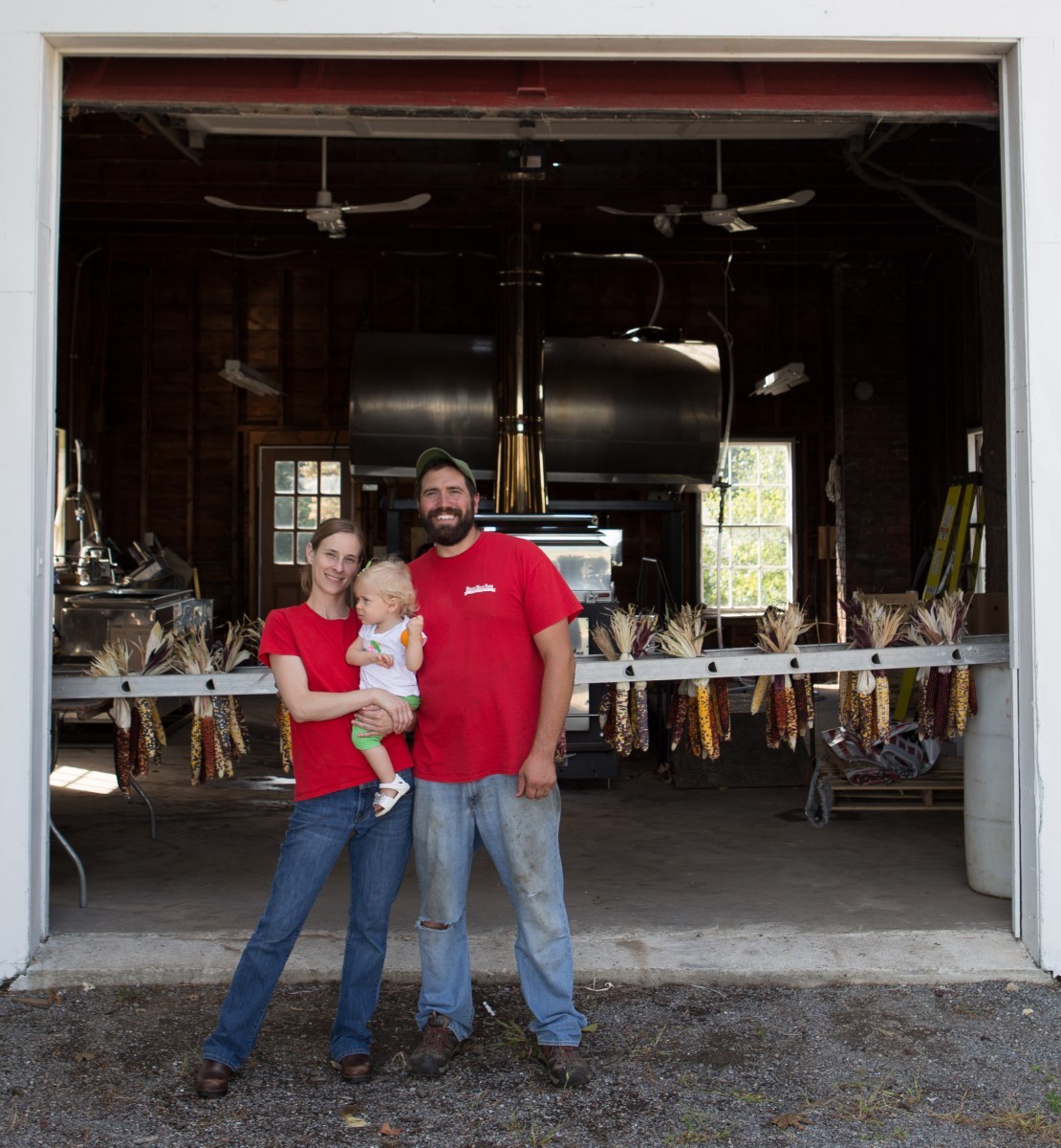 a smiling man and woman in red T-shirts, holding a small girl, stand in front of the entrance to a large barn where multi-colored ears of corn are hanging on a rack