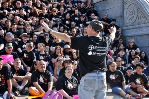 a man with raised arms faces away from the camera speaking to a large seated crowd, all wearing black T-shirts, outdoors on a sunny day