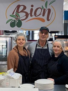 3 people in hairnets and aprons stand in front of large kitchen equipment and a sign saying Bion with an illustration of olives