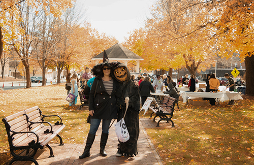 2 adults in Halloween costumes, a witch and a pumpkin-head, pose outdoors for the camera on a sunny day