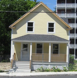 a yellow house with a gray shingled roof and a front porch with unpainted wood railings