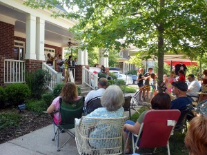a crowd of people sit in various outdoor chairs watching a band perform on a house’s front porch