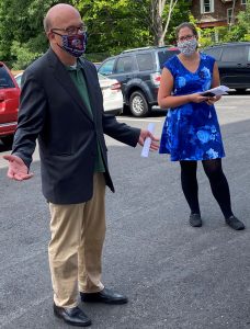 a man holding his arms out in a dark gray blazer and a woman in a blue dress stand outdoors in a parking lot, both wearing cloth face masks
