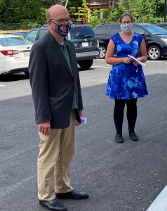 a man in a dark gray blazer and a woman in a blue dress stand outdoors in a parking lot, both wearing cloth face masks