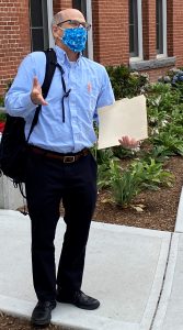 a man with glasses, a blue cloth face mask, a blue shirt, and black pants and shoes stands outdoors holding a paper file folder