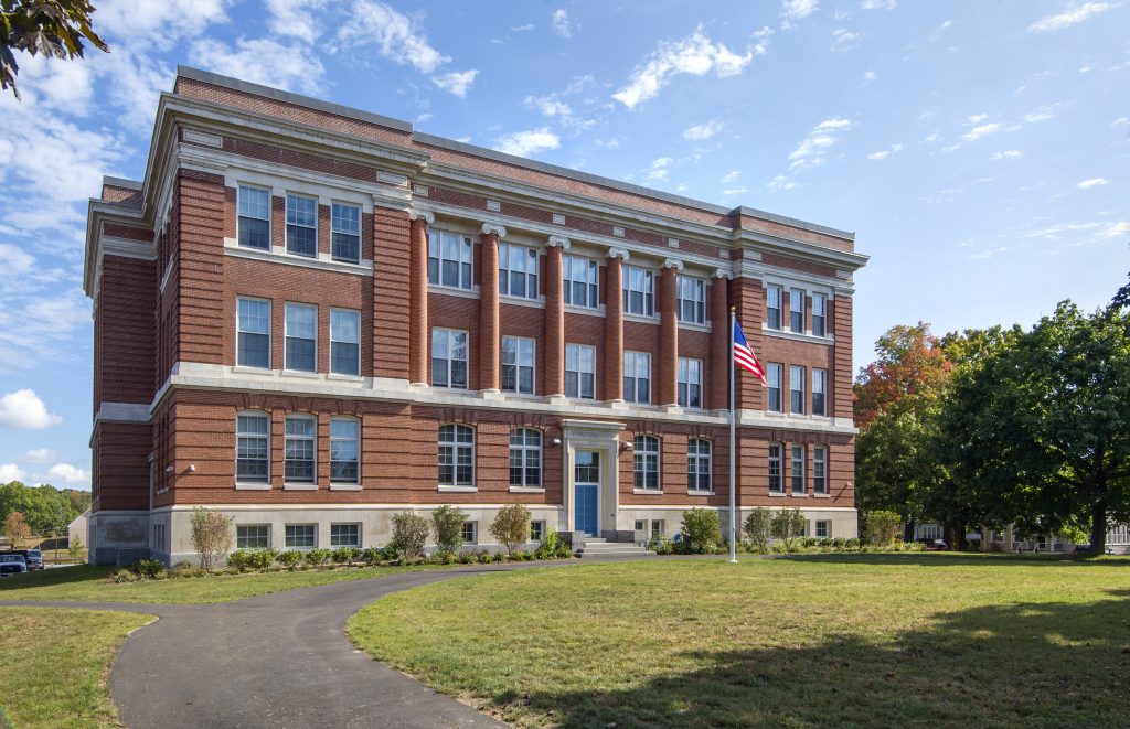 a large brick building with a curved asphalt path and grass in front, on a sunny day, with an American flag at full mast