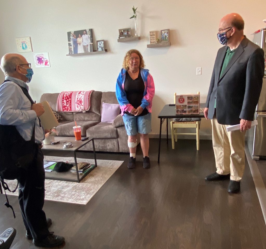 2 men and 1 woman stand indoors in an apartment with a dark wood floor and light walls, with a gray couch and photos displayed on the wall