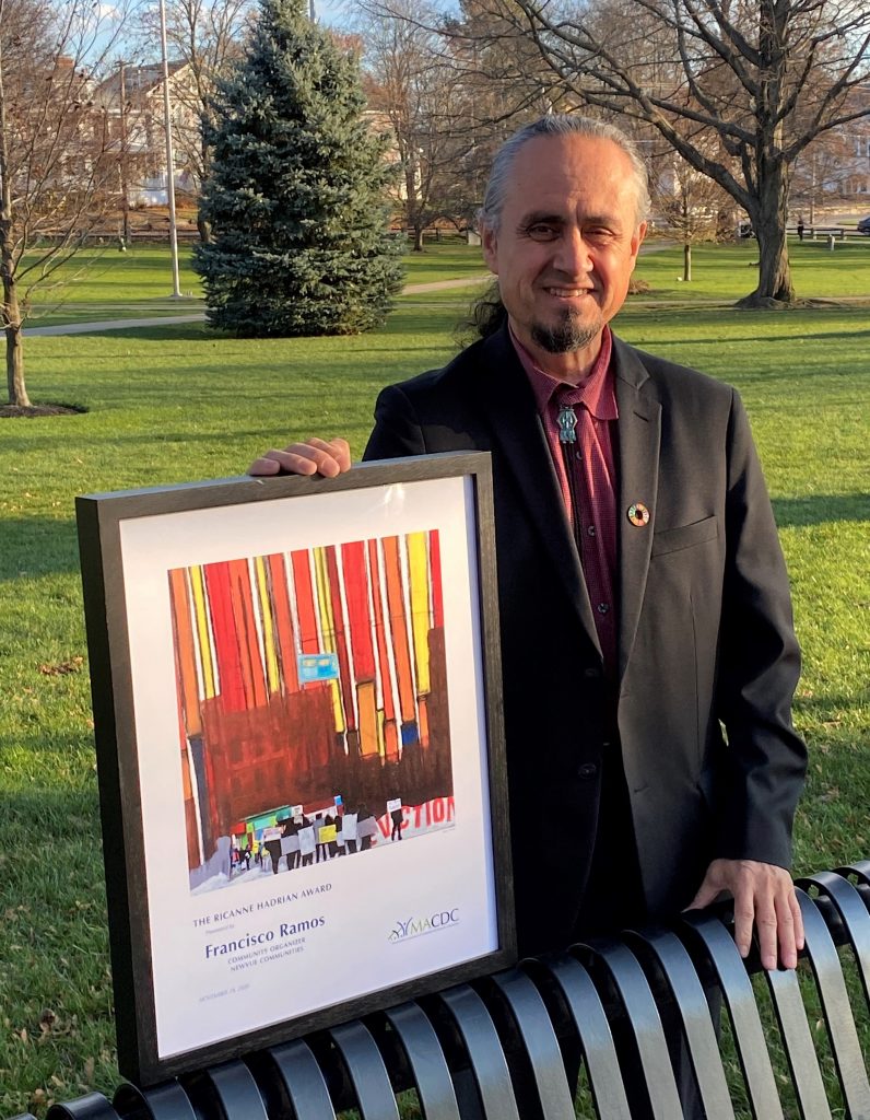 a man in a suit outdoors holding a framed award propped up on a bench, with grass and trees in the background