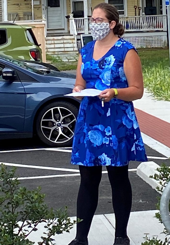 a woman in a blue dress and a white cloth face mask stands outdoors holding a piece of paper