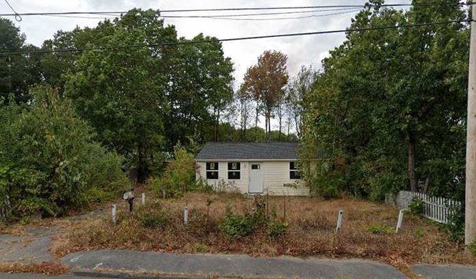 a small pale yellow house with a white door and gray roof surrounded by an overgrown lawn and large trees