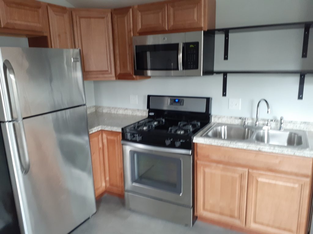 a kitchen with brown cabinets, gray marble patterned countertops, a steel double sink, steel refrigerator, gas oven, microwave oven, and open shelves