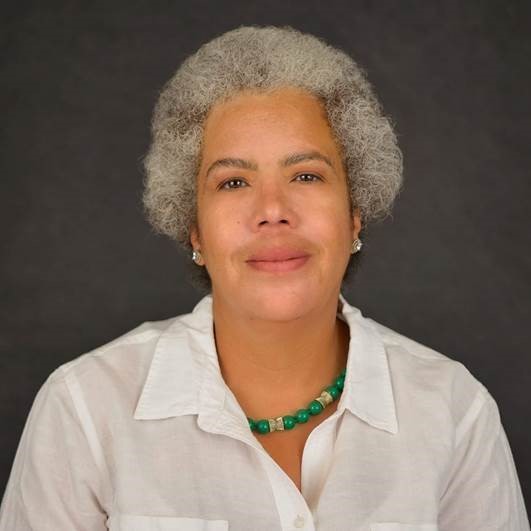 a portrait photo of a woman with curly gray hair and brown eyes, wearing a white collared shirt, green necklace, and silver earrings